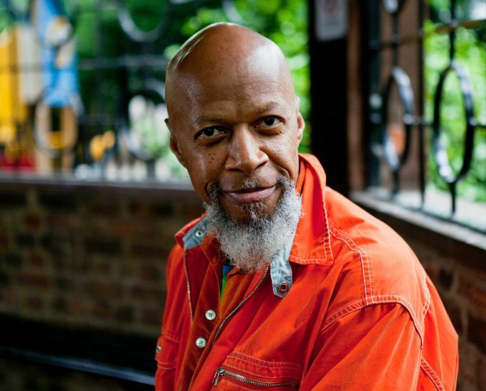 Laraaji in an enclosed outdoor space, wearing a jacket of his signature orange color, and smiling at the camera
