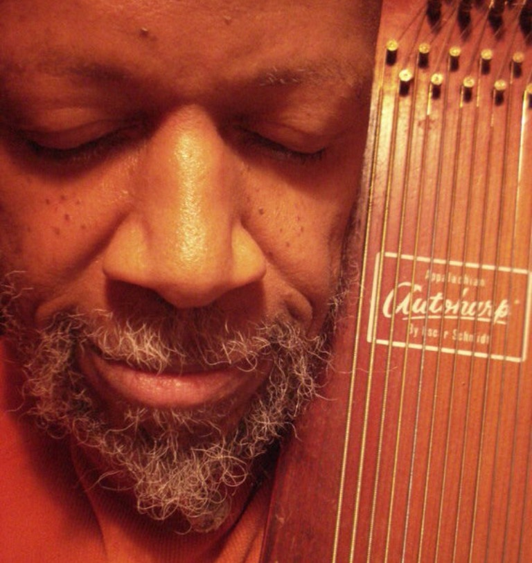 Laraaji with his eyes closed, holding his zither. 