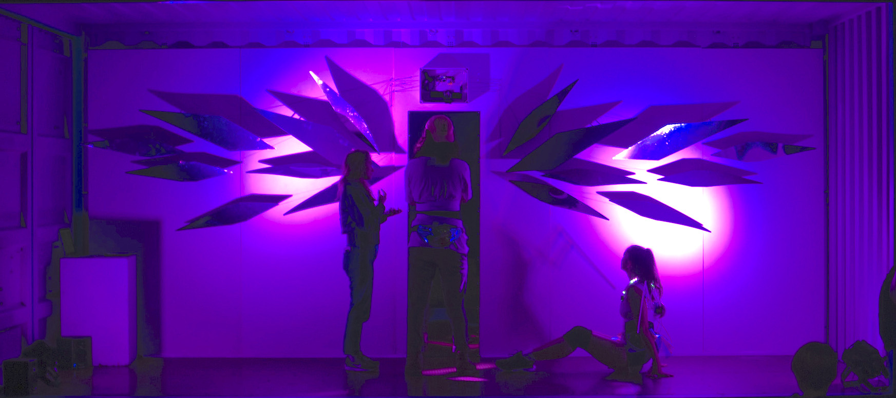 3 dancers standing on a purple lit stage with an installation of large, abstract wings made out of mirrors 