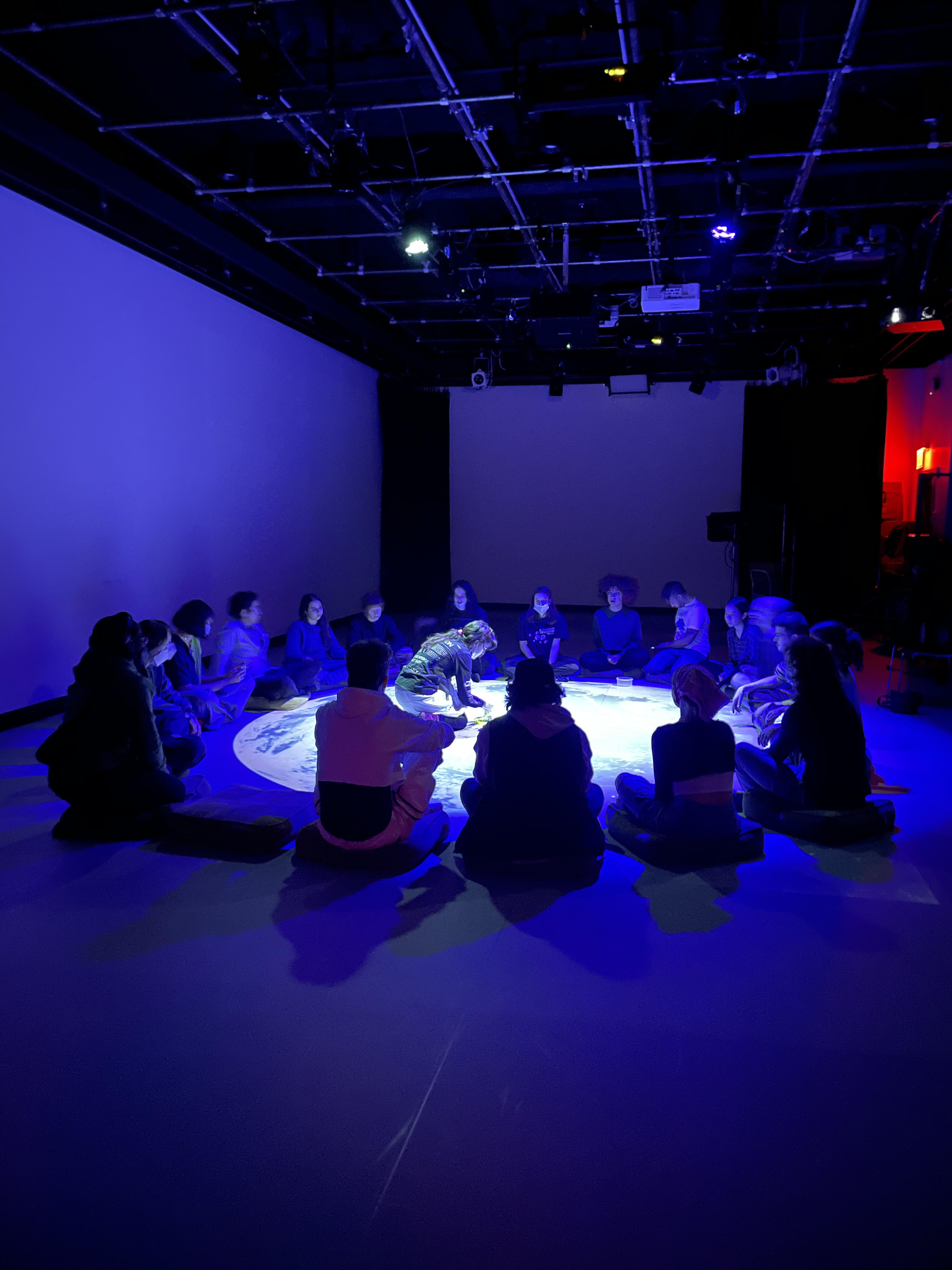 Attention Lab participants sit in a circle on the floor of the Movement Lab. A circular light is projected in the center of the group, where Amalia, the workshop facilitator, leans forward on the floor to demonstrate an exercise.