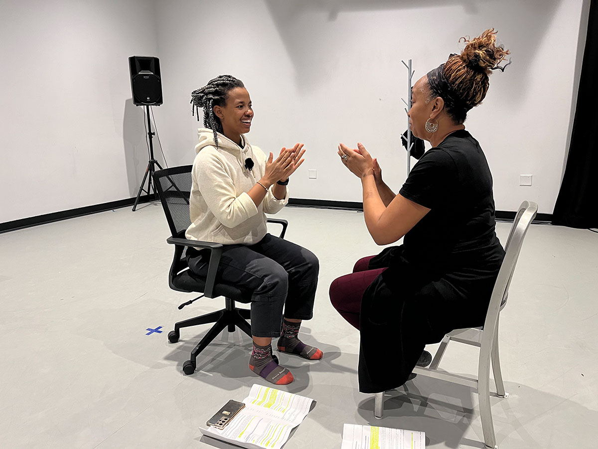 Two women sit facing each other playing a hand game