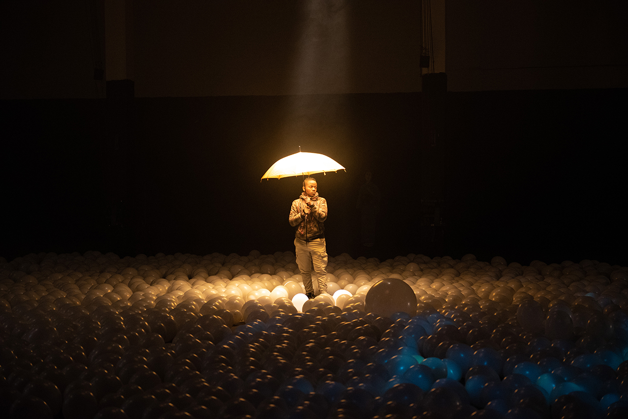A performer stands alone in a spotlight, carrying an umbrella, surrounded by balloons.