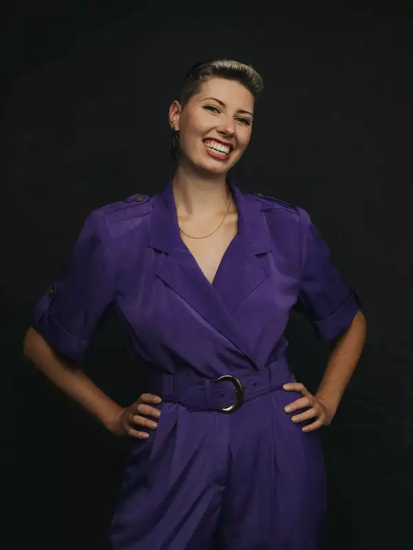 Photo of Celia standing with her hands on her hips, in a purple jumpsuit. Her hair is short and she is smiling.