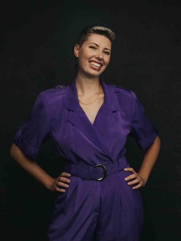 Photo of Celia standing with her hands on her hips, in a purple jumpsuit. Her hair is short and she is smiling.