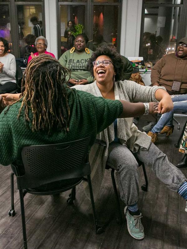 2 Black women chair dancing while others clap